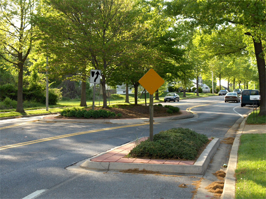 photo shows another example of a residential street 3 lanes wide, a cirle with plants fills the middle lane, and just before the cirlce from each direction the curb and grass extend in the street (a 'bulbout') for about half a lane, so that approaching vehicles must swerve to the left to miss the bulbout and then must swerve to the right to get around the circle